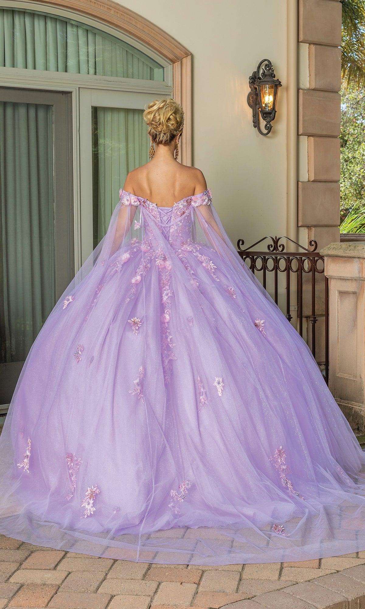 A woman with blonde hair in an updo stands outdoors with her back to the camera, wearing the Quinceanera Dress 1714 By Dancing Queen—an off-the-shoulder pastel purple ball gown adorned with floral appliqués.
